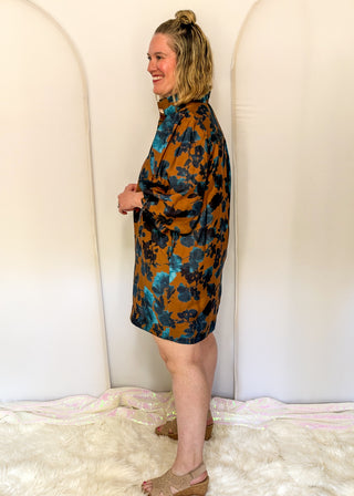 A woman wearing a short-sleeve button-up shirt dress with a blue and orange floral pattern, standing in profile against a white arched backdrop.