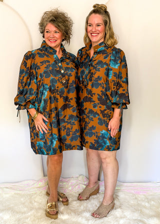 Two women modeling matching brown and teal floral mini dresses with puff sleeves and button-front collars, standing against a white wall on a white rug.
