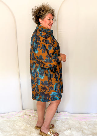 A woman modeling a high-neck, long-sleeve shift dress with a mustard orange and blue floral pattern, standing against a white arched backdrop.