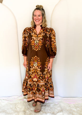 A woman wearing a brown bohemian-style maxi dress with intricate gold, orange, and red floral embroidery, standing against a white arched backdrop.