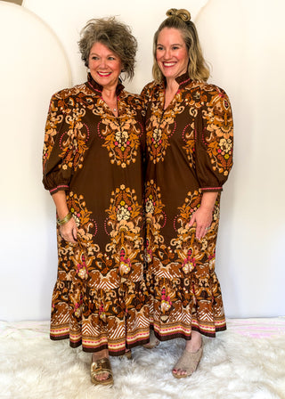 Two women standing side by side wearing matching brown bohemian-style maxi dresses with intricate gold, beige, and red floral embroidery, posing in front of a white studio backdrop.