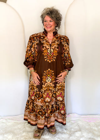 A smiling woman with short gray hair models a brown bohemian-style maxi dress with intricate gold, orange, and pink floral embroidery, standing against a white arched backdrop.