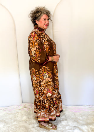 A woman modeling a brown long-sleeve maxi dress with an intricate multicolor floral and paisley print, standing against a white arched backdrop.