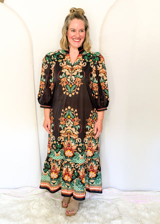 A woman modeling a long-sleeved bohemian maxi dress with a dark brown base and intricate multicolor floral and paisley patterns in green, orange, and gold, standing against a white arched studio backdrop.
