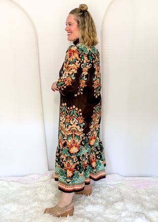 A woman wearing a long-sleeved brown velvet maxi dress with an intricate multicolor floral and botanical print, standing in a white room with white arches and a white faux fur rug.