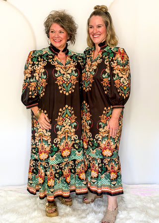Two women modeling matching long-sleeve bohemian maxi dresses with a dark brown base and vibrant multicolor floral and paisley patterns in green, orange, and gold, standing against a white studio backdrop.