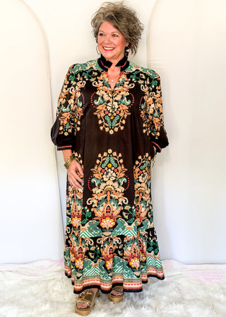 A woman modeling a long-sleeved bohemian maxi dress with a black velvet-like base and intricate multicolor floral and geometric patterns in green, orange, and gold, standing against a white studio backdrop.