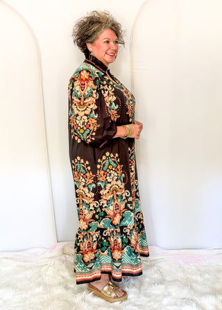 A woman modeling a long-sleeved, high-neck maxi dress with a dark brown base and an intricate, colorful traditional pattern featuring green, orange, and gold motifs, standing against a white arched backdrop.