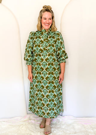 A woman modeling a high-neck, long-sleeve midi dress with a green and beige botanical pattern, standing against a white arched backdrop.