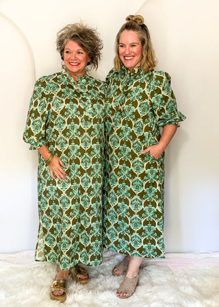 Two women modeling matching green and beige patterned maxi dresses in a bright indoor setting with a white wall and white faux fur rug.