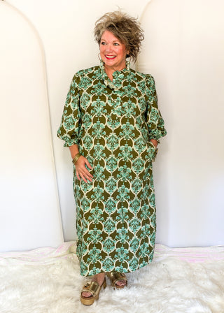 A woman wearing a green and beige patterned maxi dress with three-quarter sleeves, standing against a white arched backdrop.