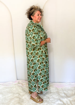 An older woman stands in profile wearing a long-sleeved button-up shirt dress with a green and beige botanical pattern, paired with gold platform sandals, set against a white arched backdrop.