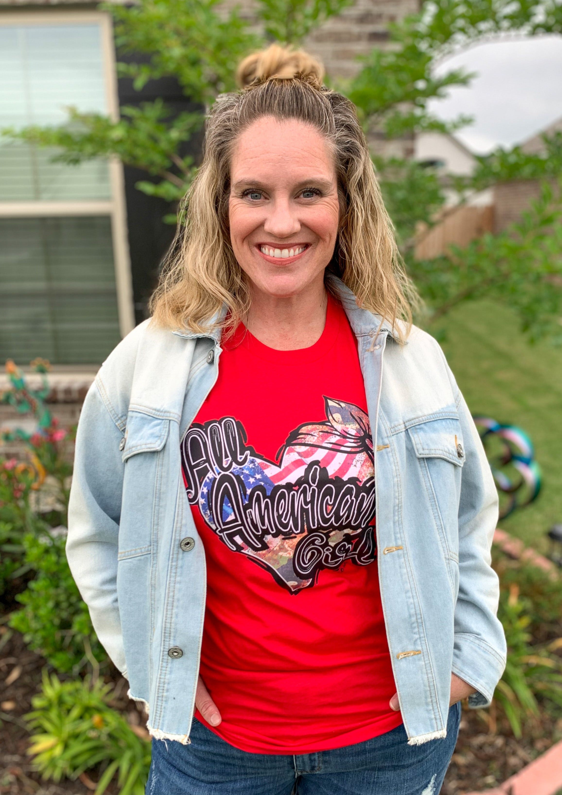 Red tee shirt with mixed pattern heart, says "All American Girl" over heart. Heart has American flag, camo, and a Rosie the Riveter headband. 