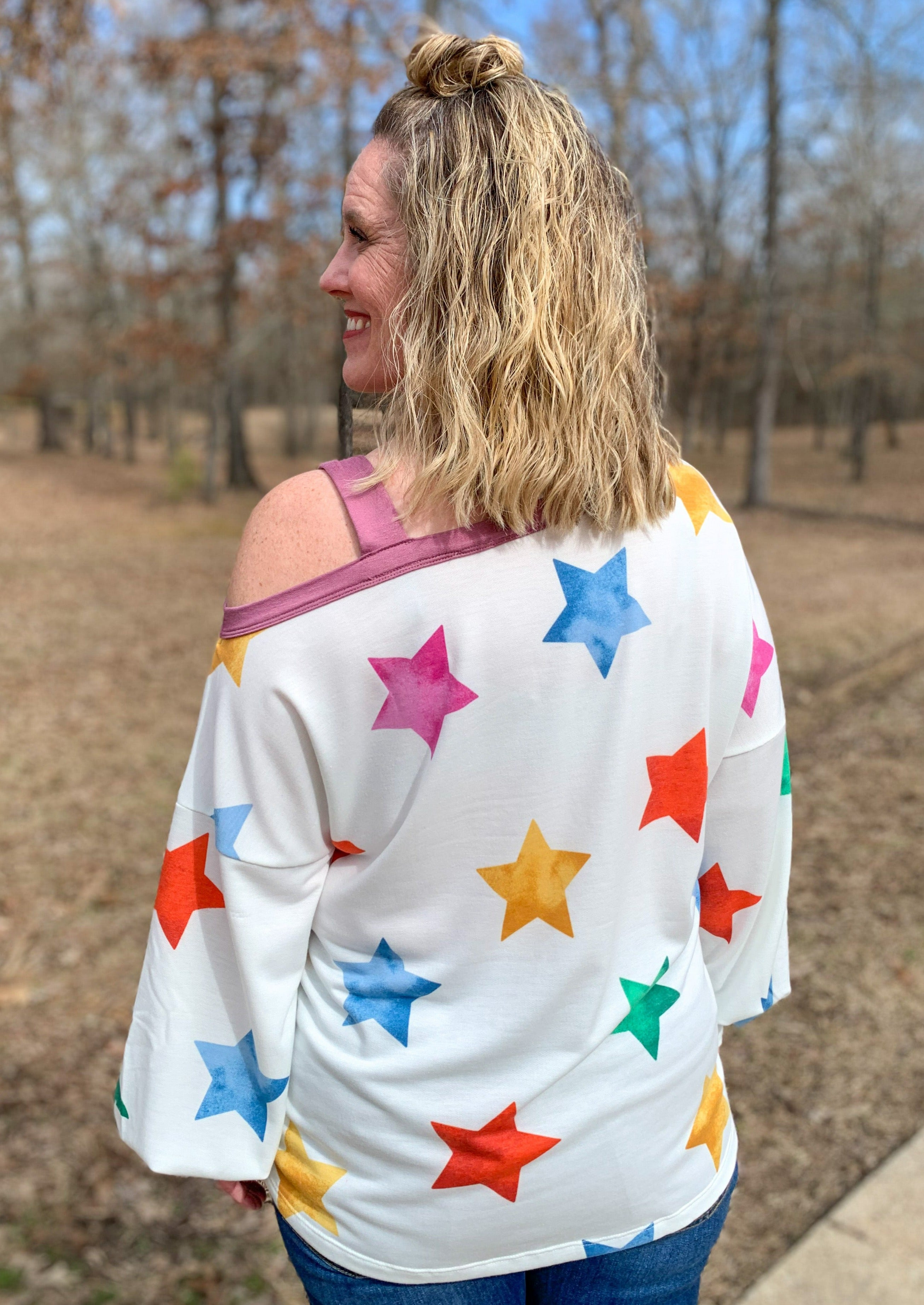 Back of White long balloon sleeve top with multicolored stars printed around/ It has a purple band at the collar and off the one shoulder with a strap on one shoulder.