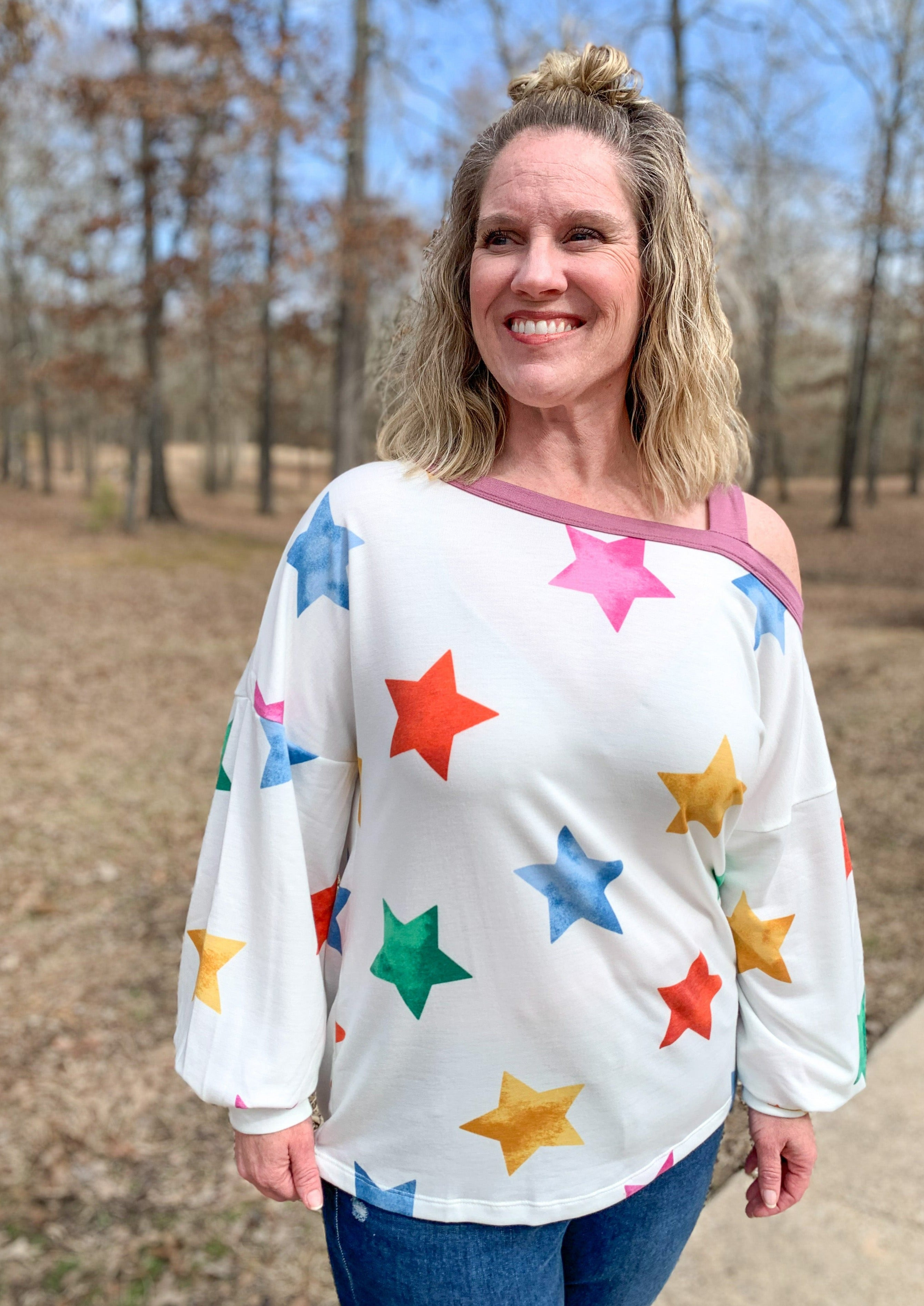 White long balloon sleeve top with multicolored stars printed around/ It has a purple band at the collar and off the one shoulder with a strap on one shoulder.