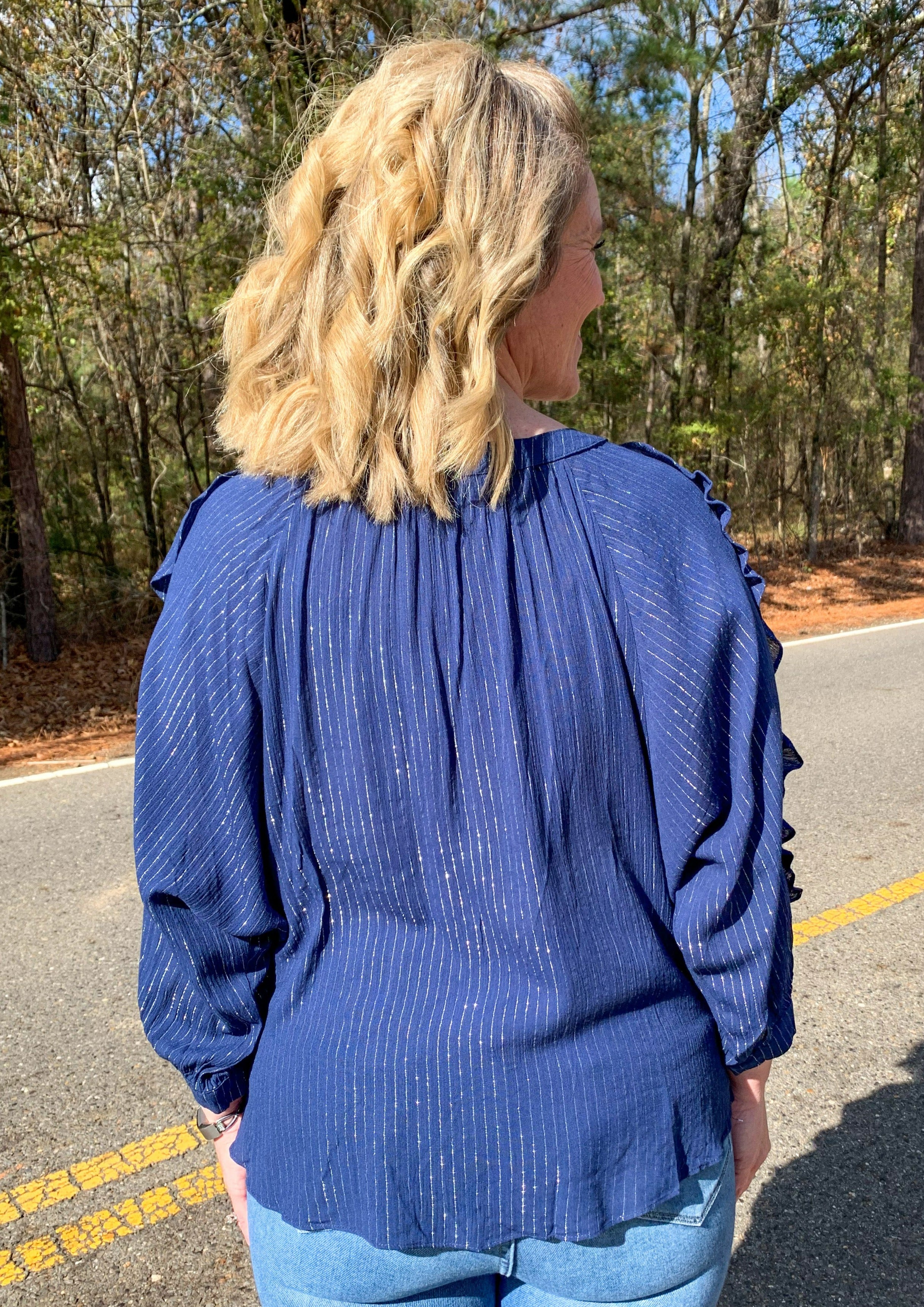 Back of Blue blouse with ruffle details on the sleeves and down the front. Has a button and small keyhole on the front. Metallic threading creates a vertical stripe pattern throughout the whole shirt. 